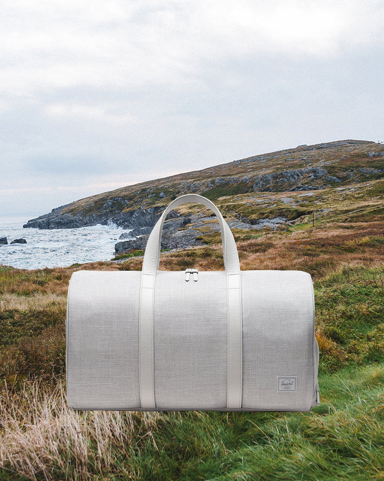 Herschel Novel Duffle in Grey Crosshatch. A grey duffle bag featuring a shoe compartment, black top handles, and a removable shoulder strap.