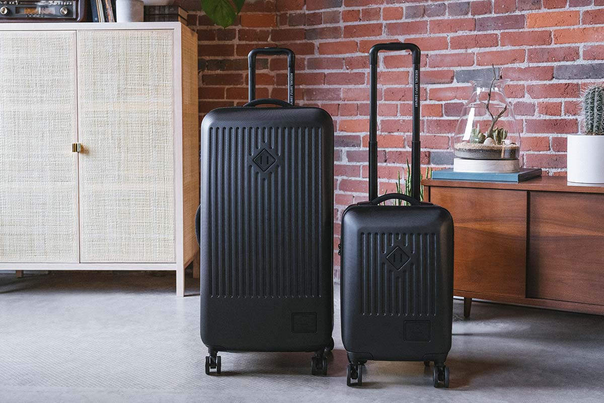 A black Trade Luggage Large & Trade Luggage Carry-On next to each other in an exposed brick apartment