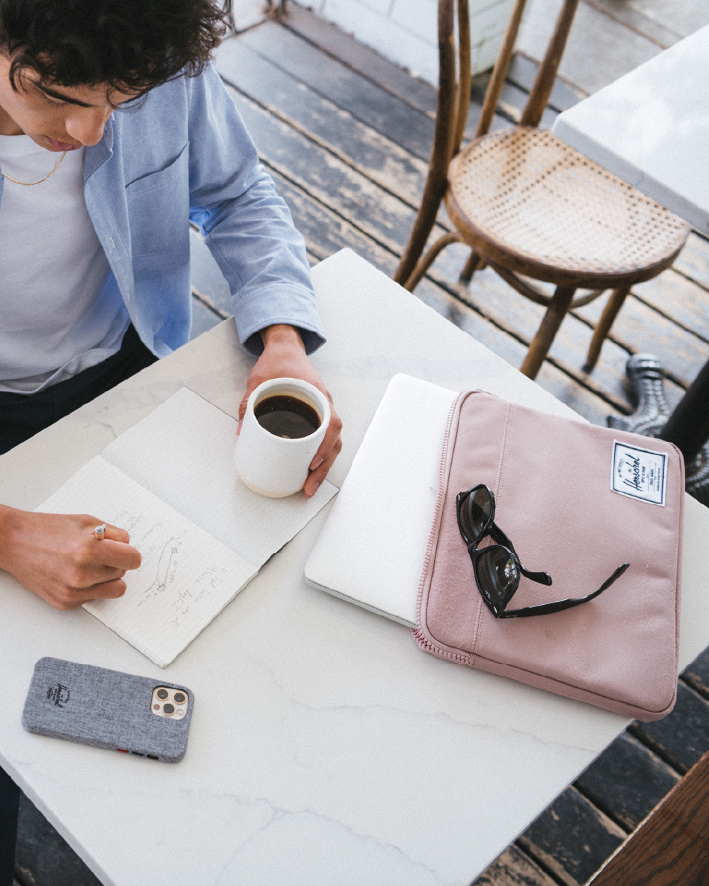 A person sitting at a cafe drinking a coffee writing in a sketchbook with their iPhone in a Herschel Classic iPhone 12 Case and a pink Anchor Laptop Sleeve on the table as well