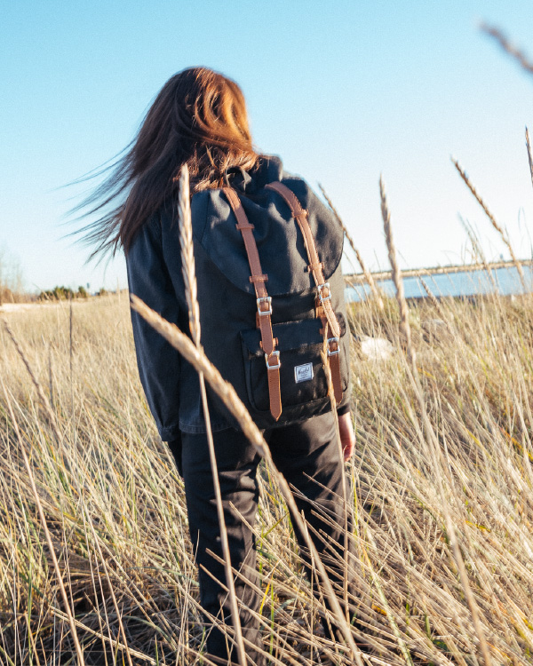 A woman walking through a wheat field carrying her Herschel Little America Backpack in Black/Tan Synthetic Leather