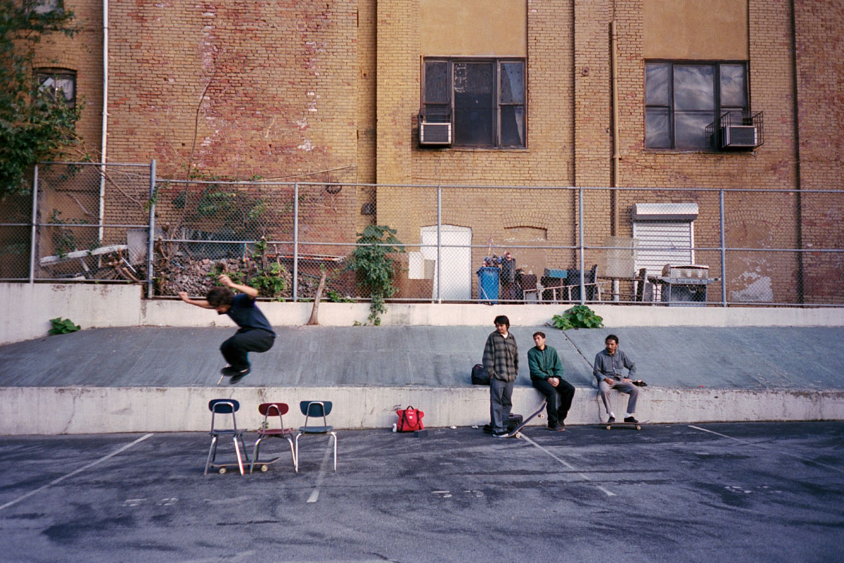 Chocolate team rider Erik Herrera hippy jumps 3 chairs in a school yard with a Herschel Supply Company x Chocolate Alexander Zip Tote in High Risk Red/Black on the ground next to Stevie Perez, Jordan Trahan, and Kenny Anderson in the background