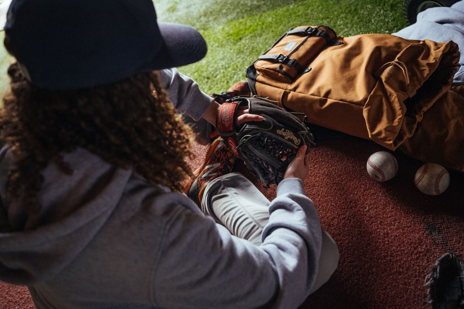 A baseball player from behind wearing their glove with their Herschel Little America Backpack Pro on the ground beside them with baseballs on the ground