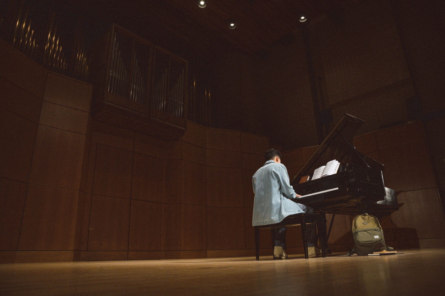 A far-away shot of a person sitting at the piano playing with their Herschel Heritage Backpack Pro on the ground beside them leaning on the piano.