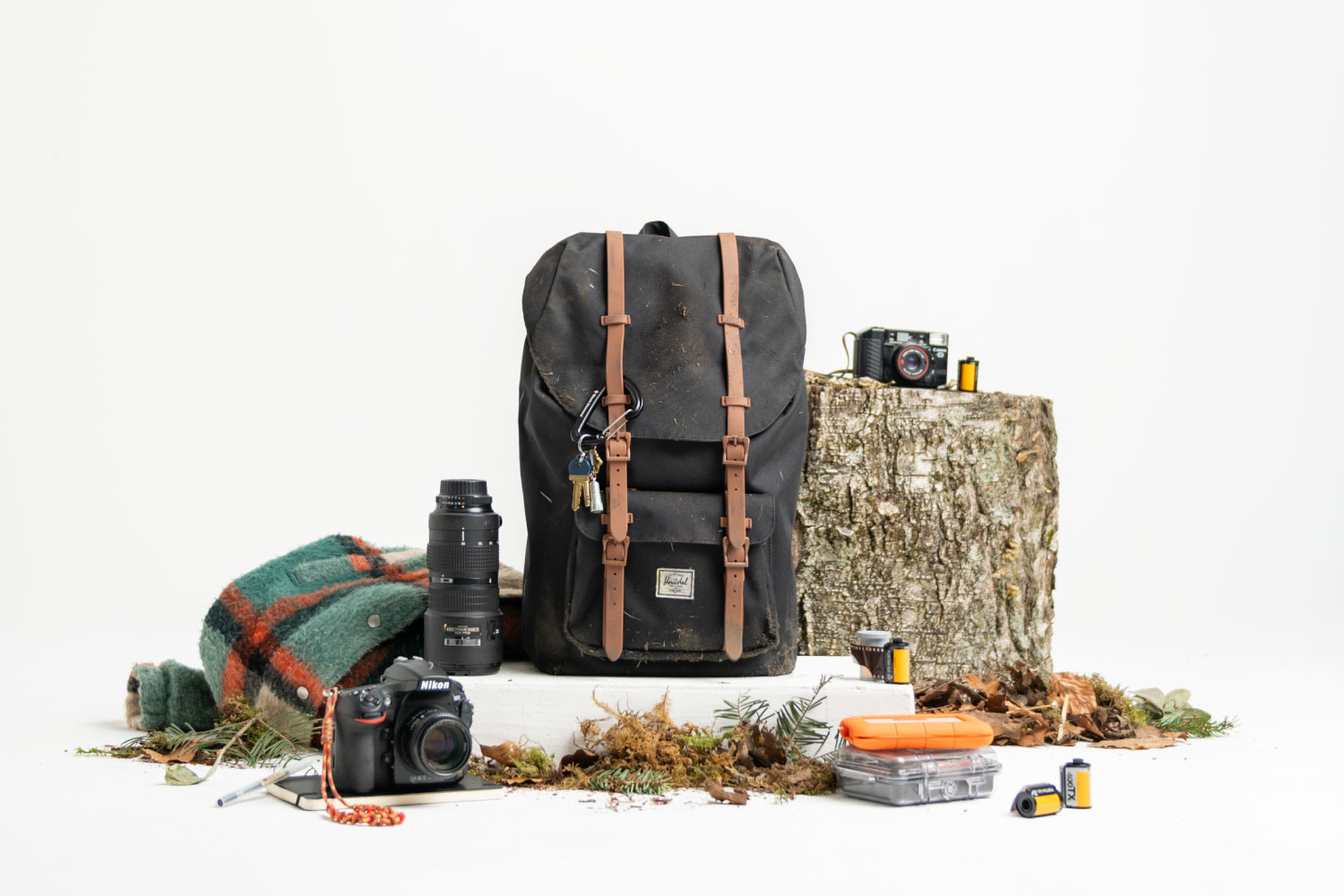 A black Herschel Little America Backpack in Black/Saddle next to a plaid flannel jacket, a camera and some film on a tree stump and an extra camera lens