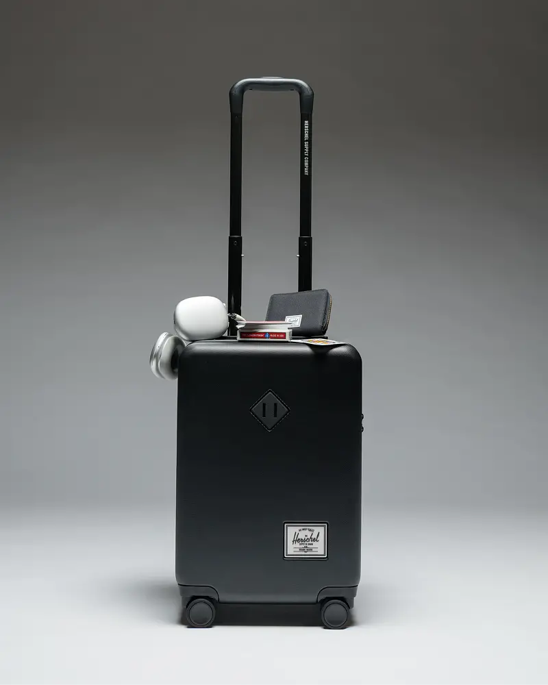 a heritage hardshell suitcase with headphones and other travel essentials sitting on top of it.